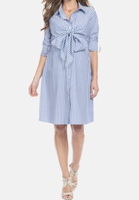 Kobieta w niebieskiej i białej pionowo paskowanej sukience koszulowej z podwiniętymi rękawami, z przodu zapinanej na guziki, z dużą kokardą zawiązaną w talii, w połączeniu z białymi sandałami na obcasie.