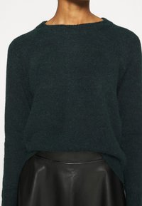 Tmavě zelený pletený svetr s volnějším střihem, kulatým výstřihem a žebrovanými manžetami, zkombinovaný s elegantní černou koženou sukní.