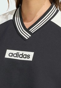 Černé atletické tričko s výstřihem do V a pruhovaným límcem; na obdélníkové nášivce je bílý logo Adidas. Hladká látka, sportovní design.