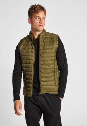 Olivgrüne Steppweste mit vertikaler Steppung, hohem Kragen und Seitentaschen, getragen über einem schwarzen Langarmshirt und dunklen Hosen.