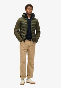 Giacca trapuntata verde con cappuccio, maniche verde oliva, maglione blu navy sotto, pantaloni khaki e sneakers neutre. Presenta un logo sulla manica.