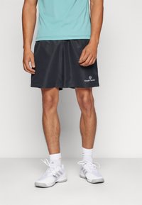 Schwarze Sportshorts mit glatter Textur und seitlichem Schlitz, mit einem weißen Logo auf der unteren linken Seite. Getragen mit einem himmelblauen Oberteil und weißen Turnschuhen.
