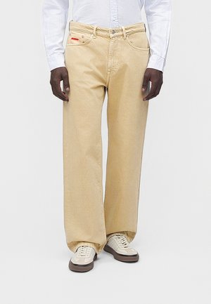 Uomo che indossa jeans beige a gamba larga, camicia bianca abbottonata e sneakers bianche, in piedi davanti a uno sfondo chiaro e uniforme.