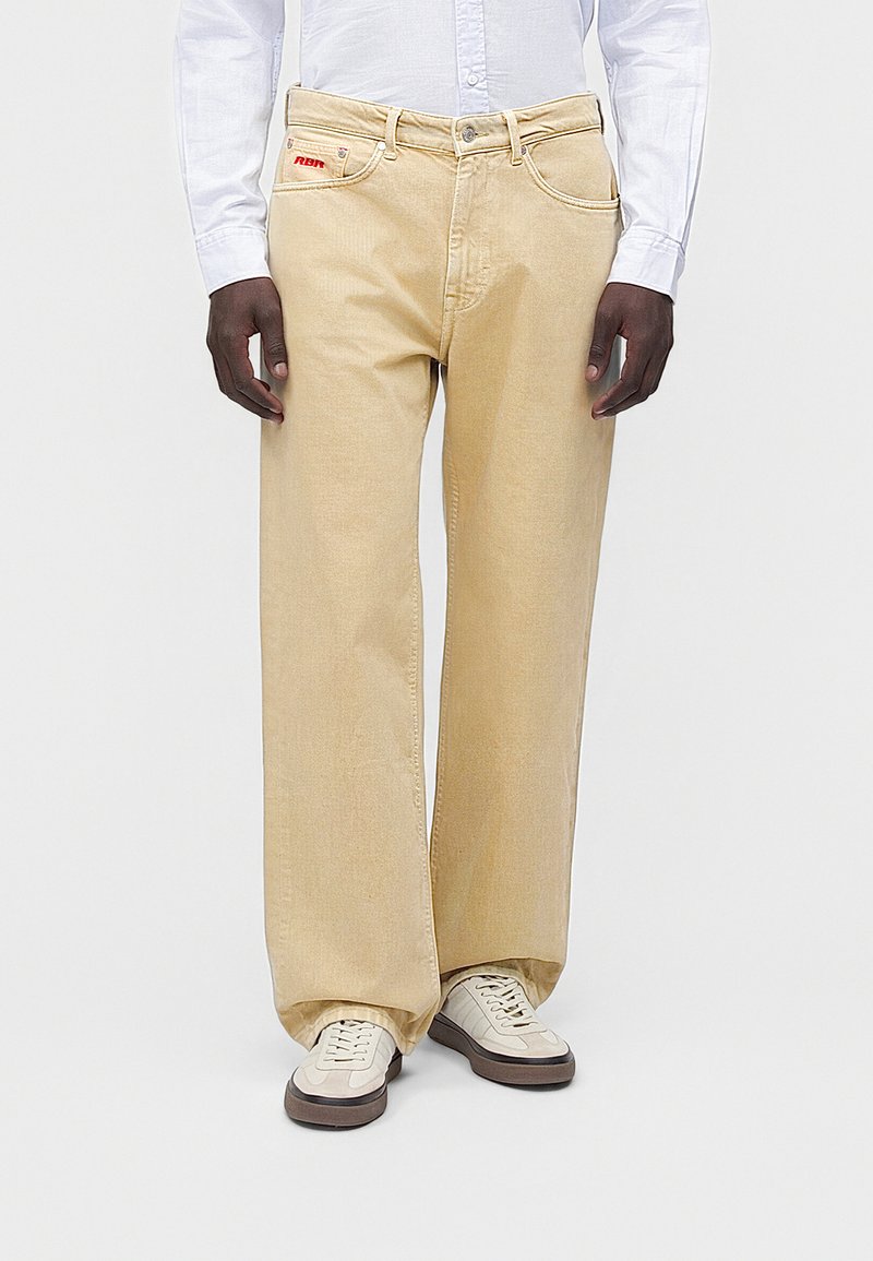 Άνδρας που φοράει μπεζ τζιν παντελόνι φαρδύ, λευκό πουκάμισο με κουμπιά και λευκά αθλητικά παπούτσια, στέκεται μπροστά σε ένα απλό φωτεινό φόντο.