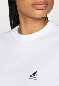 Valge puuvillane T-särk, millel on must Kangol logo koos känguru graafikaga ja tekst "KANGOL J.LINDEBERG" vasakul rinnal.