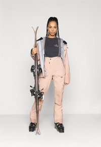 Conjunto de esqui com uma jaqueta impermeável rosa claro e overall combinando, com botas de esqui pretas e um par de esquis, em fundo simples.