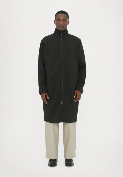 Black long coat with high collar and full zipper, made of textured material. Paired with light beige pants and black shoes.