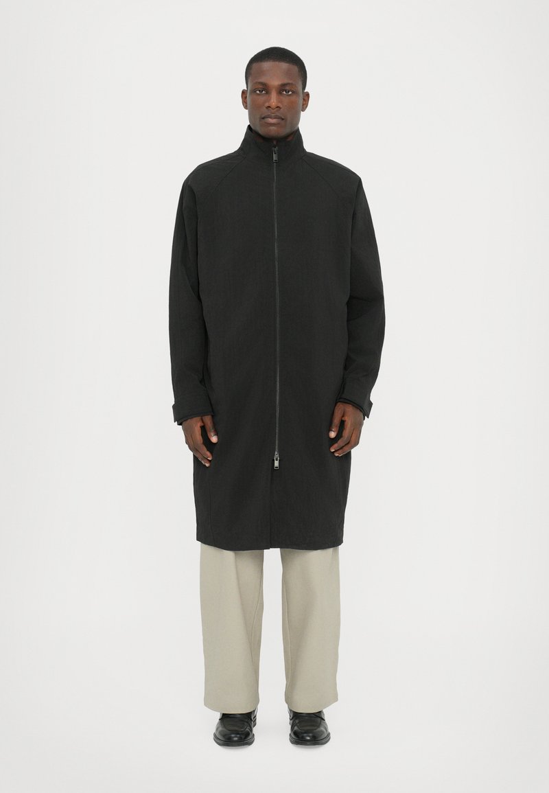 Black long coat with high collar and full zipper, made of textured material. Paired with light beige pants and black shoes.