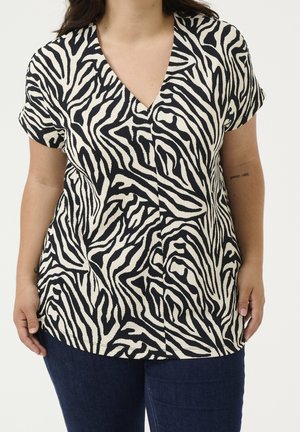 Frau trägt eine kurzärmelige, schwarz-weiße V-Ausschnitt-Bluse mit Zebramuster und dunkelblaue Jeans, steht vor einem schlichten hellen Hintergrund.