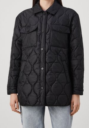 Sort quiltet jakke med bølgemønster, knaplukning foran, klaplommer på brystet, båret over lyseblå jeans.