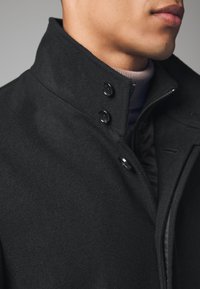 Zwarte wollen jas met een hoge kraag, voorzien van twee zwarte knopen en een rits. Gladde textuur, op maat gesneden ontwerp en minimalistische details.