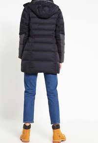 Zwarte donsjas met capuchon en grijze stoffen mouwen, met een doorgestikt ontwerp. Gecombineerd met een blauwe spijkerbroek en beige enkellaarzen.