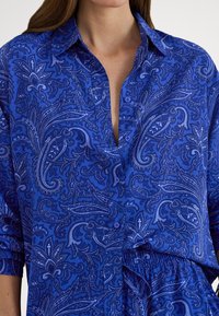 Modrá košeľa na gombíky s paisley vzorom v svetlejších modrých odtieňoch, s vyhrnutými rukávmi a voľným strihom. Hladká textúra látky.