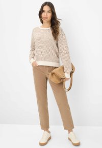Maglione crema con motivo a diamante marrone, pantaloni in corduroy beige e una borsa in suede color cammello. Sneakers bianche con dettagli color cammello.