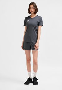 Ung kvinna stående i mörkgrå sportkläder med shorts, vita strumpor och svarta träningsskor mot en enfärgad vit bakgrund.