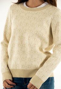 Suéter beige de tejido suave con un patrón repetido de formas "Z" de color claro. Cuello y puños acanalados con sutiles toques de brillo.