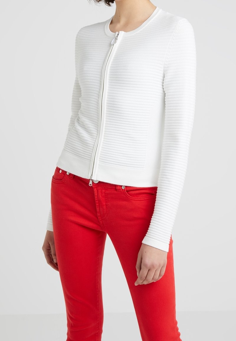 Personne portant un haut blanc côtelé à manches longues avec fermeture éclair et un pantalon ajusté rouge vif sur un fond gris clair.