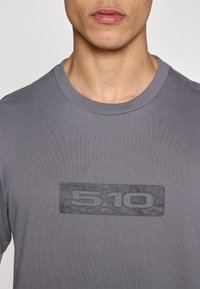 Γκρι μπλούζα με κοντά μανίκια και στρογγυλή λαιμόκοψη, με ανάγλυφο ελαστικό λογότυπο που φέρει τους αριθμούς "5.10" στο κέντρο του στήθους.