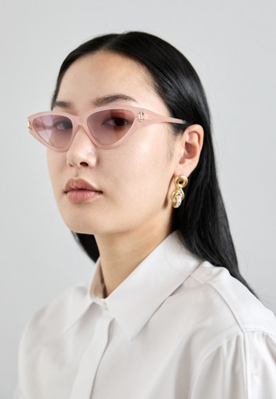 Cat-eye sunglasses with light pink frames and tinted lenses, featuring side hardware accents. Model wears a white button-up shirt.