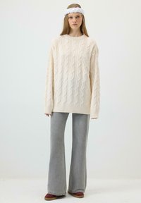 Noor naine pikkade juustega, seljas kreemikas kaabelpitsist suuri kampsunit, hallid laia säärega püksid, bordoopunased kingad ja valge peapael.