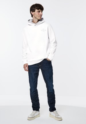 Felpa bianca con cappuccio e foderatura grigia, indossata con jeans blu scuro e sneakers bianche, caratterizzata da un tessuto testurizzato e una vestibilità rilassata.