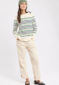 Femme portant un bonnet en tricot vert, un pull crème rayé de vert et de bleu, un pantalon beige ample et des chaussures plates noires, debout les mains dans les poches.