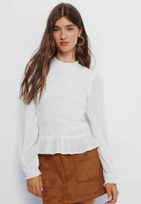 Bílá blůza s dlouhým rukávem, textured smocking a volánkovým spodním okrajem. Má vysoký límec a nařasené rukávy, doplněné hnědou semišovou sukní.