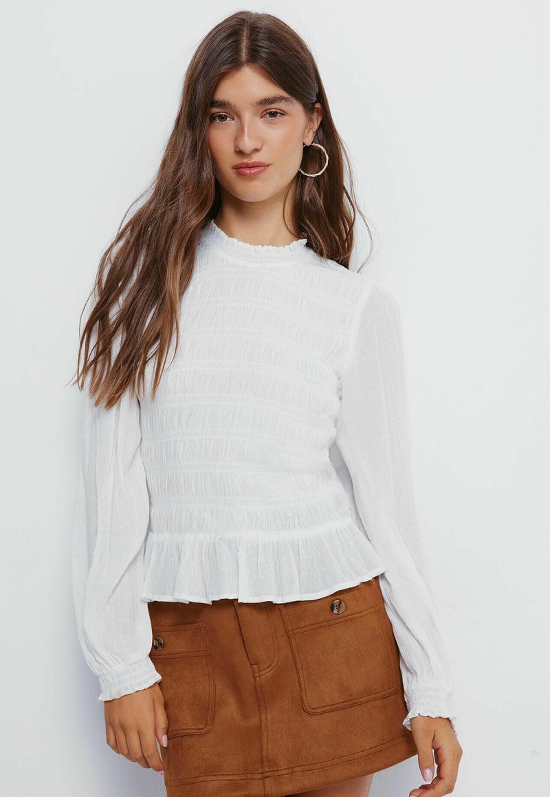 Bílá blůza s dlouhým rukávem, textured smocking a volánkovým spodním okrajem. Má vysoký límec a nařasené rukávy, doplněné hnědou semišovou sukní.