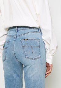 Personne portant un jean taille haute bleu clair avec poches arrière et une chemise blanche à manches longues, main reposant près de la hanche.