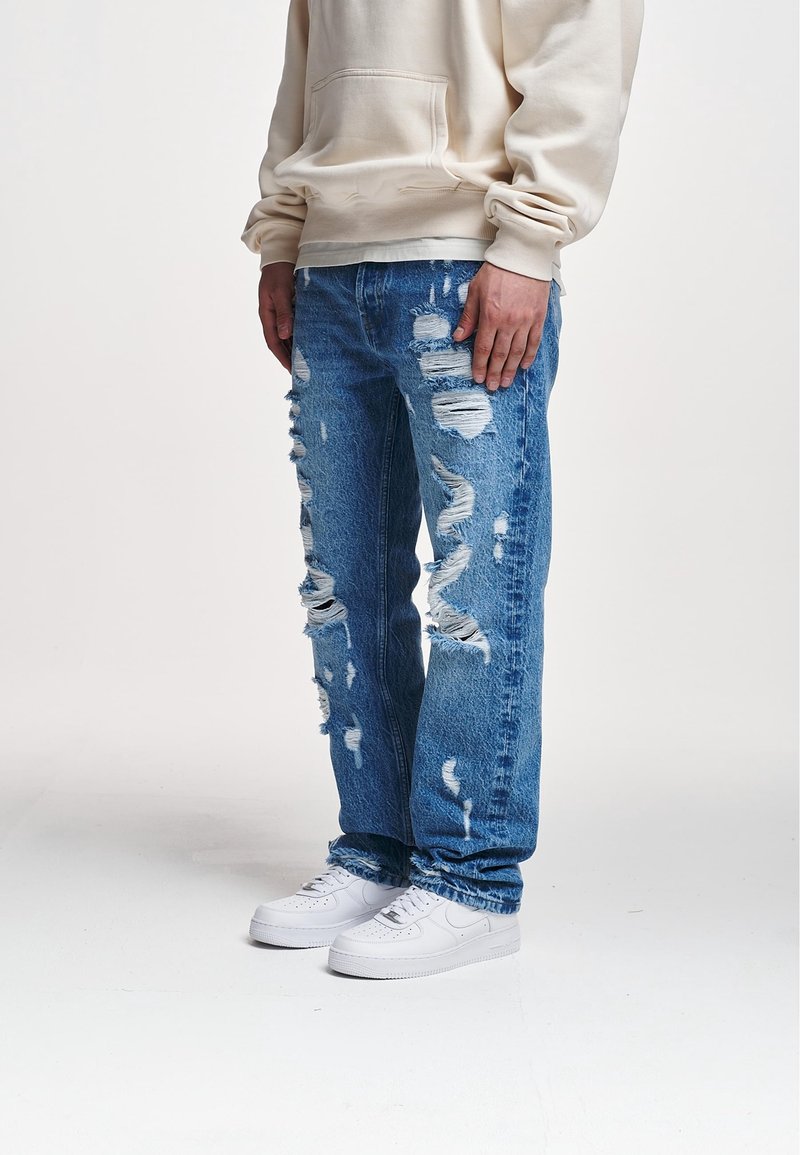 Personne portant un sweat à capuche beige, un jean bleu très déchiré et des baskets blanches, debout devant un fond clair uni.