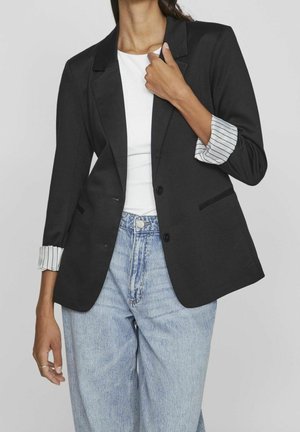 Kobieta ubrana w czarną marynarkę z podwiniętymi pasiastymi mankietami, białą bluzkę i jasnoniebieskie jeansy z wysokim stanem na jednolitym tle.