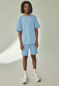 Vaaleansininen puuvillainen t-paita, jossa on rento istuvuus ja lyhyet hihat, yhdistetty samaan sarjaan kuuluvien shortsien kanssa. Pehmeästä materiaalista, käytetty valkoisten tennareiden kanssa.