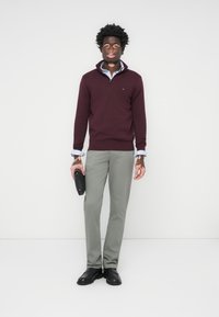 Bordeaux trui met een ritskraag, grijze-tailored broek en zwarte schoenen. Model houdt een zwart object vast, met zichtbaar gekleurde blauwe manchetten.
