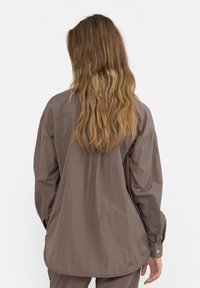 Osoba s dlouhými vlnitými blond vlasy, oblečená v nadměrné hnědé košili na knoflíky a odpovídajících kalhotách, stojící na pozadí s jednoduchým bílým pozadím.