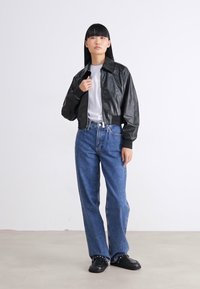 Kvinne som står iført en svart skinnjakke over en hvit skjorte, blå høy i livet jeans, og svarte sko mot en ensfarget hvit bakgrunn.