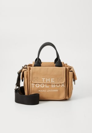 Bolso de herramientas de lona beige con acentos de cuero negro, que cuenta con asas superiores, correa ajustable y texto frontal: "THE TOOL BOX MARC JACOBS."