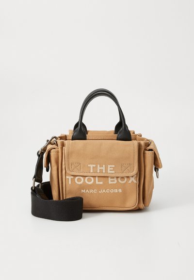 Sac à outils en toile beige avec des accents en cuir noir, doté de poignées supérieures, d'une bandoulière réglable et d'un texte à l'avant : "THE TOOL BOX MARC JACOBS."