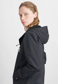 Μαύρο αδιάβροχο jacket με κουκούλα, μπροστινό φερμουάρ και πλευρικές τσέπες. Διαθέτει λογότυπο και ομαλή υφή εξωτερικά.