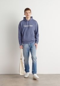 Felpa con cappuccio blu navy con logo bianco "Calvin Klein", abbinata a jeans azzurri e scarpe da ginnastica bianche. Tiene una borsa tote color crema.
