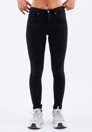 Jean noir slim porté par une personne avec le ventre découvert, mains tenant la ceinture, associé à des baskets blanches de sport.