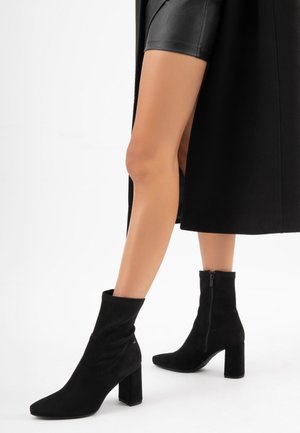 Stivaletti neri in suede con punta a forma di cono, tacco a blocco, asta elasticizzata e dettaglio con zip laterale, caratterizzati da una texture liscia e linee di design sottili.