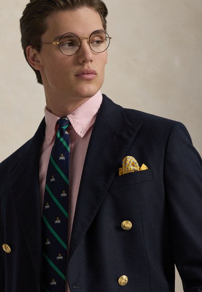 Jonge man met ronde bril, een marineblauwe dubbelgeknoopte blazer met gouden knopen, roze overhemd, gestreepte stropdas en een geel pochet kijkt opzij.