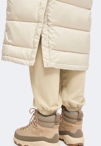 Μπεζ παλτό puffer πάνω από κρεμ παντελόνι jogger και καφέ μποτάκια πεζοπορίας με ροζ κορδόνια σε απλό ανοιχτό φόντο.