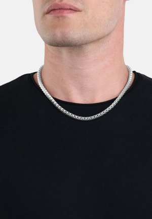 Collana in argento con un design rotondo e liscio, posata su una maglietta nera a girocollo. Realizzata in metallo con una finitura lucida.