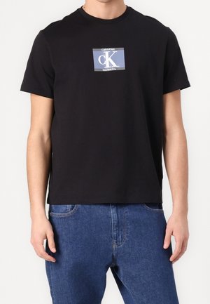 Person wearing a black Calvin Klein t-shirt with blue and white logo on chest, paired with blue denim jeans.