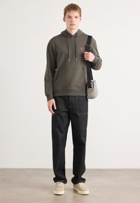 Felpa verde scuro con tasca frontale e logo rosa, abbinata a jeans neri a gamba larga e sneakers beige. Inclusa borsa a spalla casual.