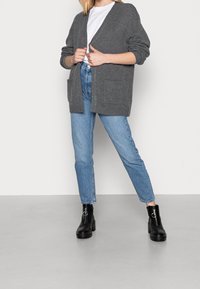 Grauer übergroßer Cardigan mit strukturiertem Strick, offener Vorderseite und zwei Fronttaschen, kombiniert mit hellblauen Jeans und schwarzen Ankle-Boots.