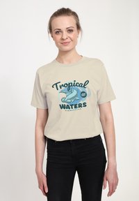 Krémové bavlněné tričko s grafikou dvou žraloků kladivounů a nápisem "Tropické vody" v modrozelené a černé typografii.