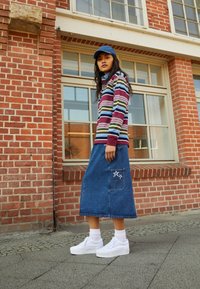 Pasiasta koszula z długim rękawem w kolorach czerwonym, zielonym i niebieskim; dżinsowa midi spódnica z naszywką w kształcie gwiazdki; białe trampki na platformie; niebieska czapka; tło z czerwonej cegły.