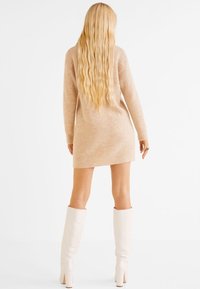 Abito in maglia beige con maniche lunghe, al di sopra del ginocchio, in tessuto testurizzato, e stivali bianchi al ginocchio. Capelli lunghi e mossi visibili da dietro.